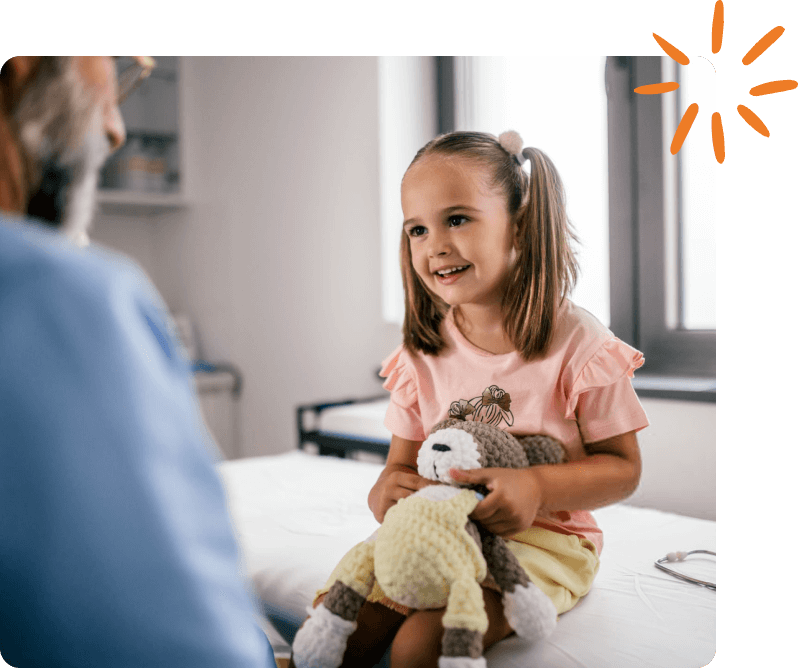 Young child holding a stuffed animal while smiling at a healthcare provider during a pediatric visit.