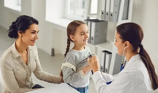 Pediatrician listens to a smiling child’s heartbeat while a parent looks on.