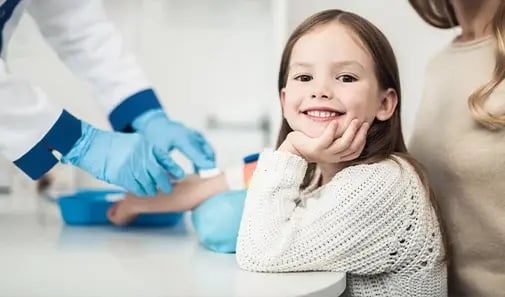 Smiling child sits with a parent while a healthcare provider prepares for a blood draw.