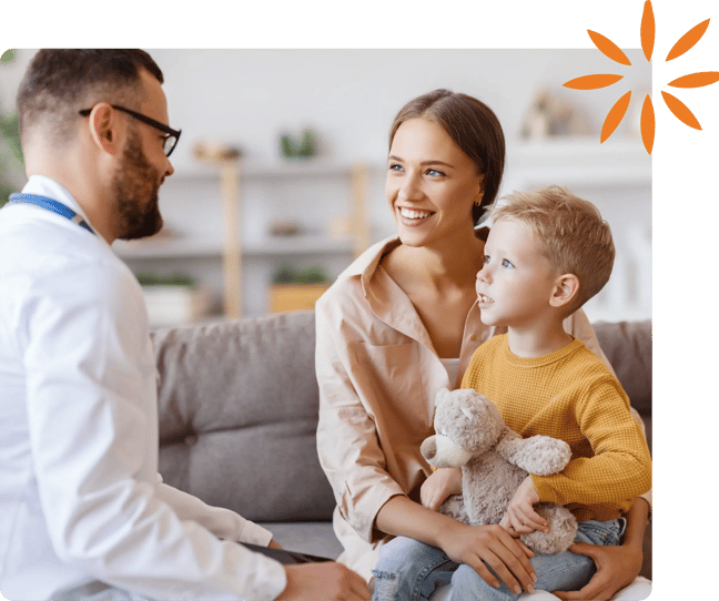 Pediatrician talks with a parent and young child holding a stuffed animal during a visit.