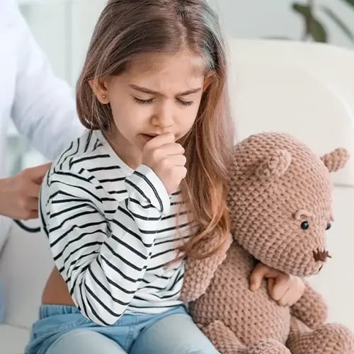 Young child coughs while holding a stuffed animal during a pediatric visit.
