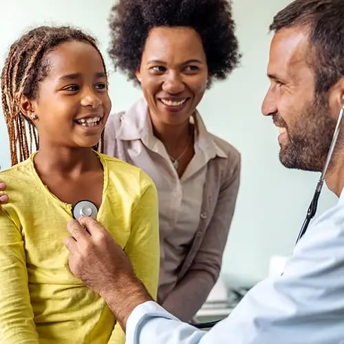 Pediatrician listens to a smiling child’s heartbeat while a parent watches.